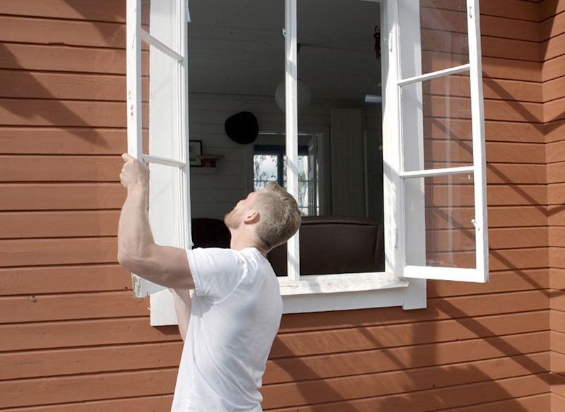 Man i vit t-shirt öppnar vitt fönster på röd träfasad, glimt av ljust vardagsrum.