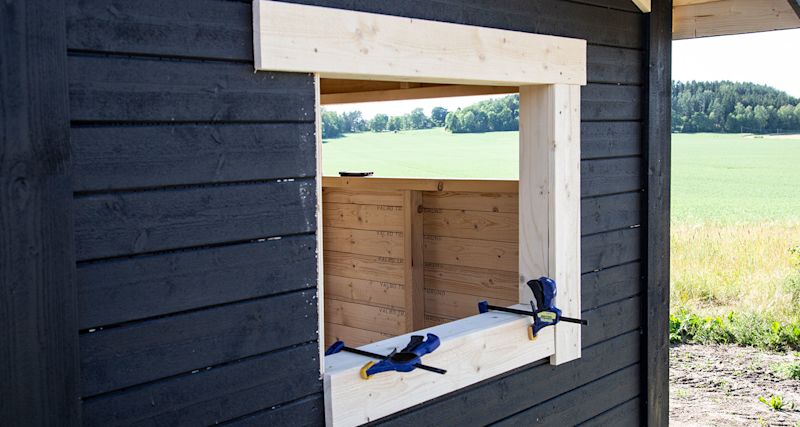 Omålat träfönster i svart trävägg med blå-gula klämmor, utsikt över grönt fält