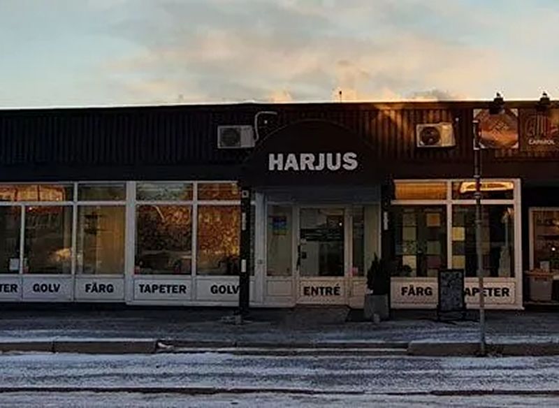 En butik med svart fasad och stora glasfönster, där skylten ovanför entrén visar namnet HARJUS och fönstren annonserar produkter som golv, färg och tapeter.