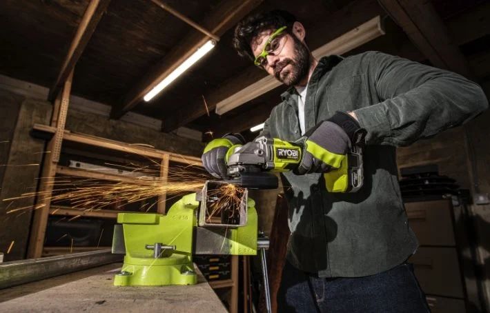 Person slipar metall i grönt skruvstäd med grön Ryobi-vinkelslip, gnistor flyger i mörkt garage.