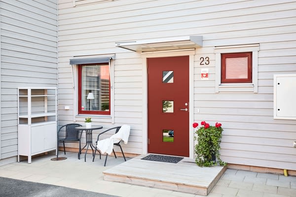 Vit träfasad med röd ytterdörr och små fönster, svarta utemöbler, vita hyllor och röda rosor.