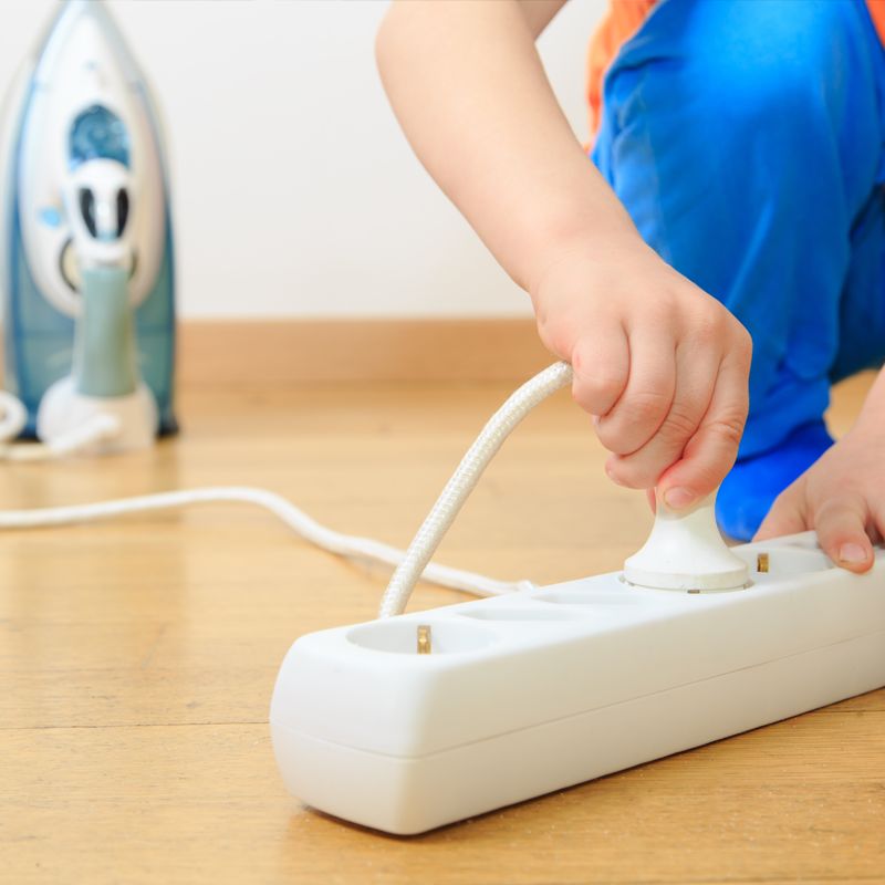 Ett barn drar ur elkontakt ur vit grenuttag på trägolv, strykjärn i bakgrunden