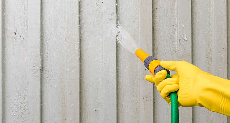 Hand i gul gummihandske som sprutar vatten med grön slang mot grå träpanel