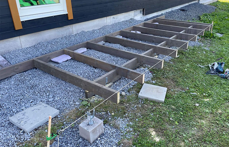 Träram för altandäck på grått krossat grus under svart träfasad, med grön gräsmatta.