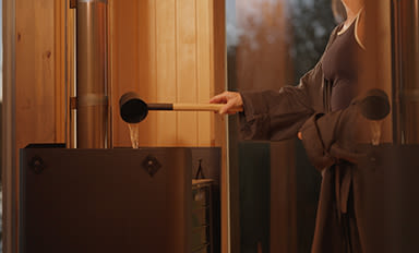 Person öser vatten med träskopa på het stenkamin i varm träbastu.