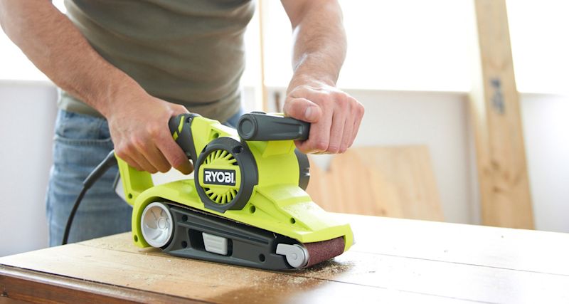 Två händer slipar träyta inomhus med neongrön Ryobi-bandslip och sågspån.