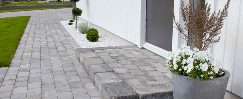 Vit husfasad, grå stenläggning, prydnadsgrus, små gröna buskar och kruka med vita blommor