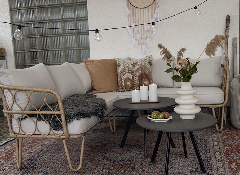 Bohemisk uteplats med rottingsoffa, beige och bruna kuddar, svarta bord med ljus och vas