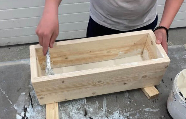 Person rör om vit spackel i rå trälåda med spackelspade på arbetsbänk.