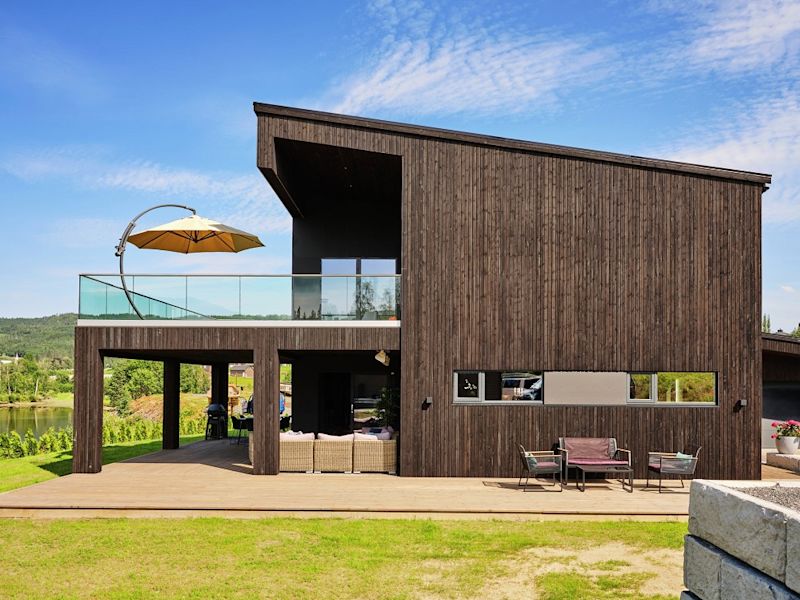 Modernt tvåvånings brunt trähus med glasräcke, gul parasoll på balkong och utemöbler.