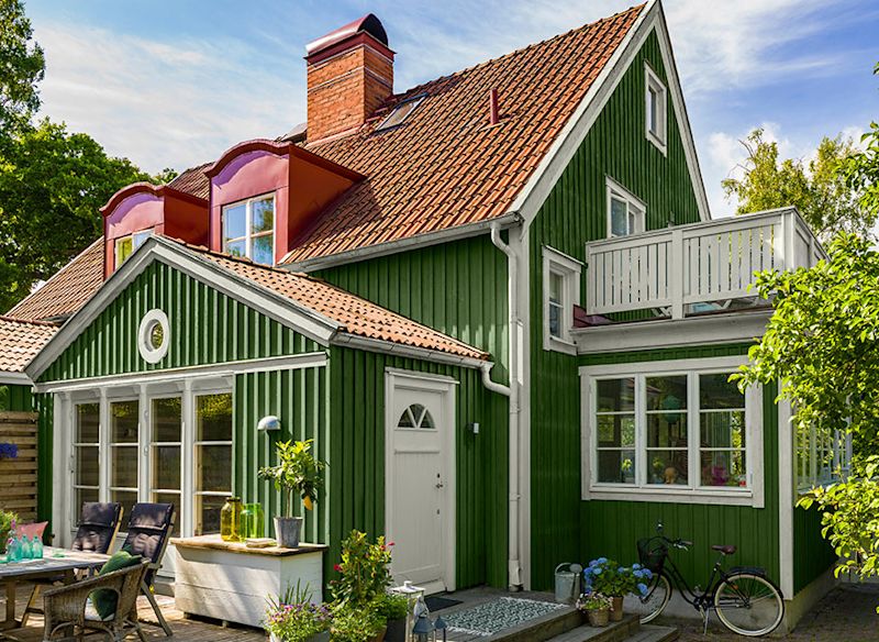 Grönt trähus med vitt fönster och dörr, rött tegeltak, balkong, uteplats med möbler och cykel.