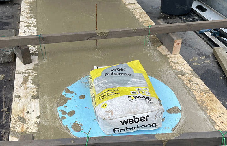 Vit-gul påse med Weber finbetong på blå skyddsfilm omgiven av nyslaget grått cement på träställning.