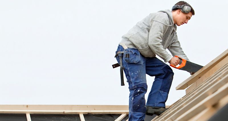 Byggarbetare i grå luvtröja och blå arbetsbyxor sågar träbjälkar på takstomme.