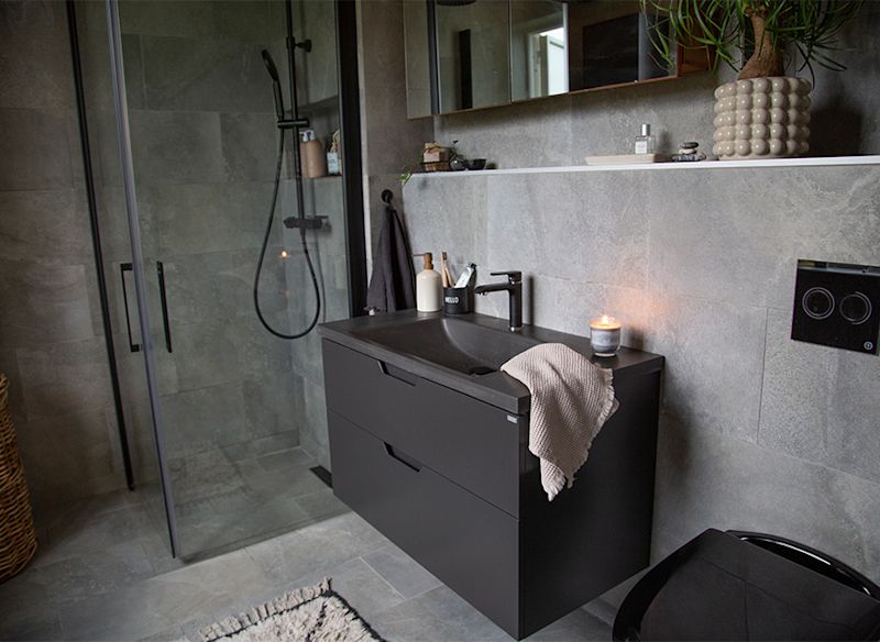 Modernt grått kaklat badrum med svart vägghängt handfat, glasdusch och tända ljus.