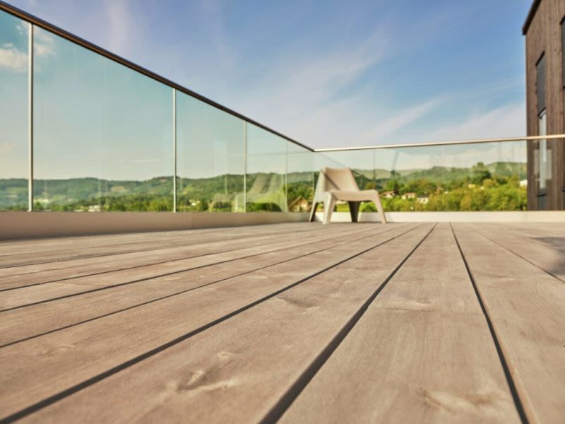 Rymlig träterrass med glasräcke, vit stol, grön skogshorisont och blå himmel