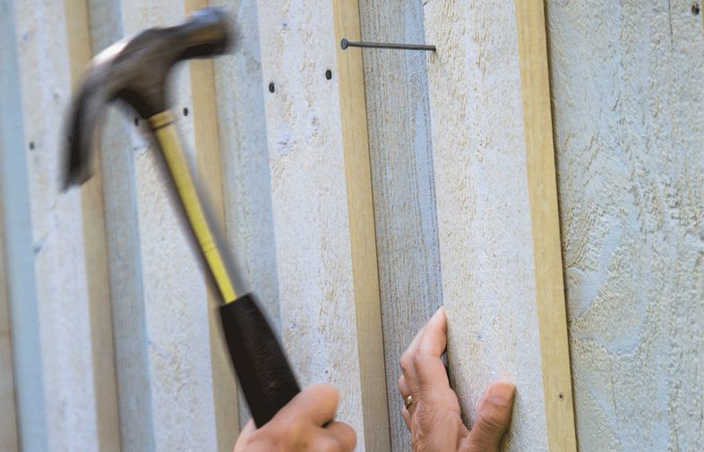 Närbild: hammare slår spik i ljusgrå träpanel med en hand som håller bräda.
