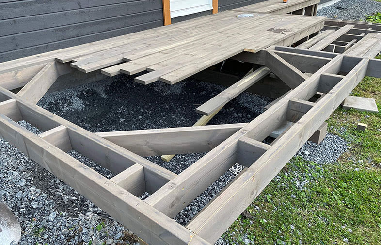 Oavslutat grått trädäck med synliga reglar och liggande plankor över svart grus vid husfasad