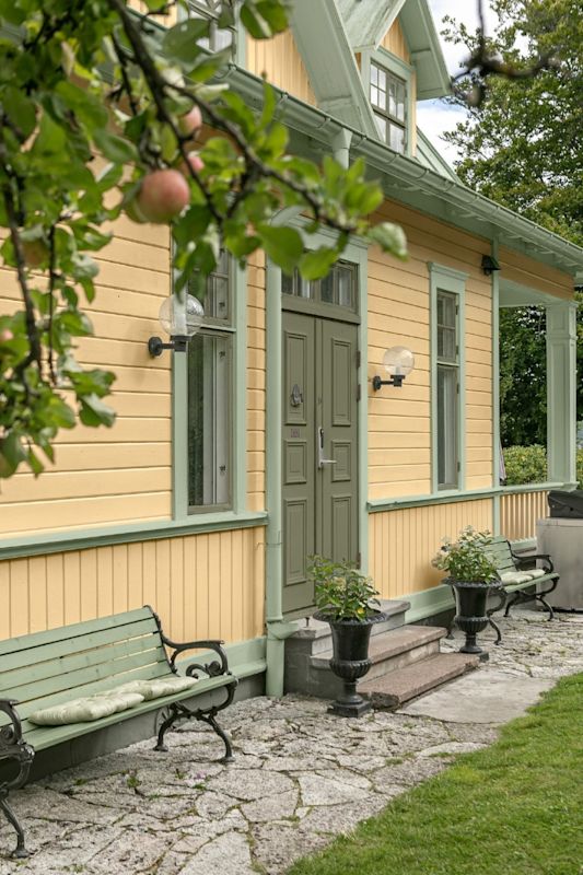 Gul trävilla med grön dörr och fönsterbågar, bänkar och blomkrukor under äppelgren.