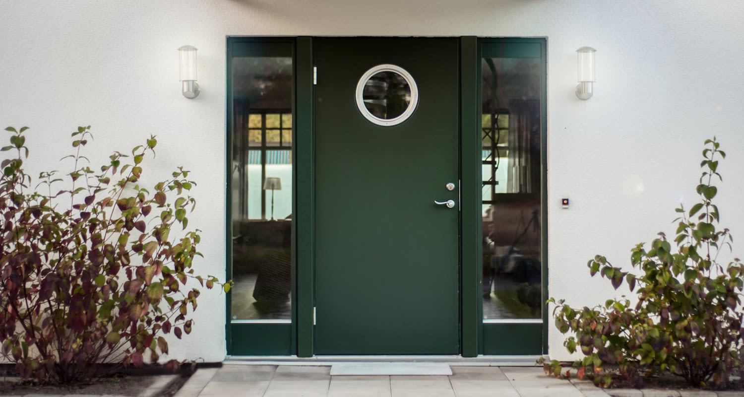 Mörkgrön entrédörr med runt fönster, glaspartier, vit fasad, vägglampor och höstfärgade buskar