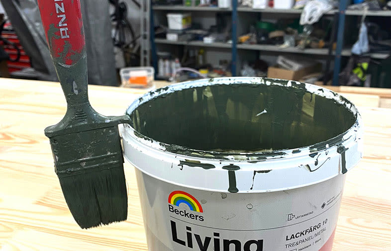 Beckers Living-färgburk med mörkgrön lack och pensel, verkstadshyllor i bakgrunden.