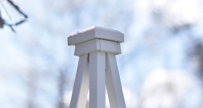 Vit dekorativ träpelare med fyrsidig pyramidtopp mot blå himmel.