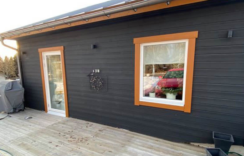 Svart trähusfasad med orange fönster- och dörrkarmar, träaltan och röd bil reflekteras.