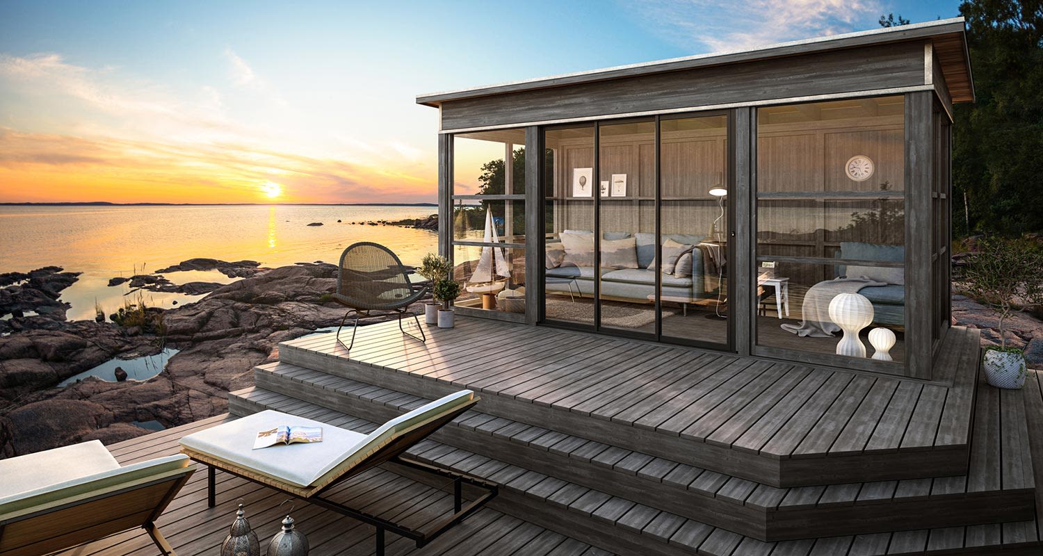 Glasväggad trästuga på klippig kust vid solnedgång, trädäck med solsängar och stol.