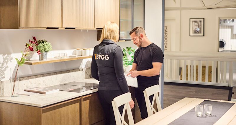 Två personer diskuterar ritning i ljus köksvisning med träskåp, marmorbänk och gröna växter.