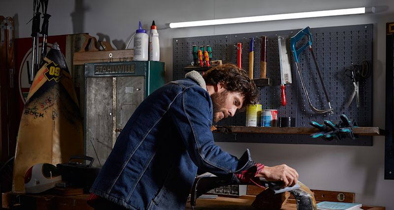 Person i blå jeansjacka filar träbit på träbänk i verkstad med grå verktygstavla.