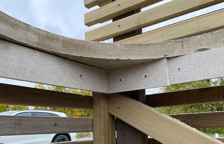 Detalj av utomhusbänk med grå betongsits och ljusbruna trälameller mot grönska och vit bil