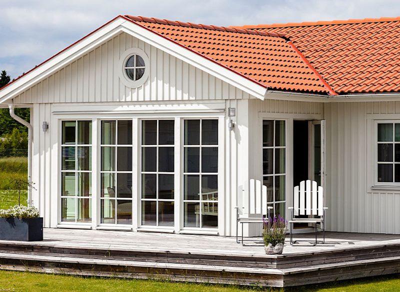 Vit trävilla med rött tegeltak, stora glasdörrar mot altan med vita stolar och blommor.