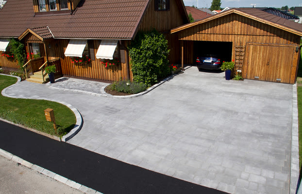 Brunt trähus med vita markiser, röda blommor i fönster, grå uppfart och öppet garage med svart bil.