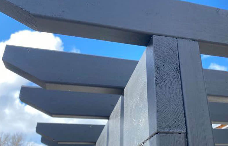 Närbild på mörkgrå pergolabjälkar mot blå himmel och vita moln.