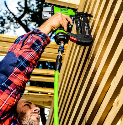 Person i rutig skjorta spikar ljusa träplankor med grönsvart tryckluftsspikpistol mot blå himmel