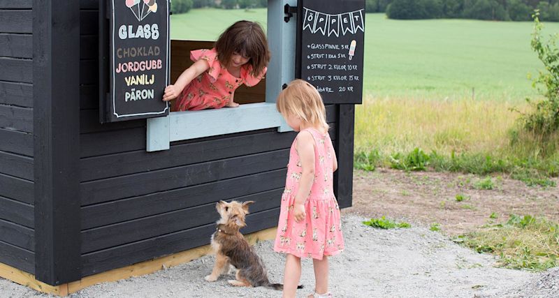Två flickor i rosa klänningar och en liten hund vid svart glasskiosk med färgglada griffeltavlor