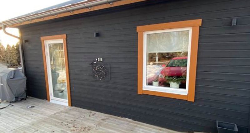 Svartgrå träfasad med orange karmar runt glasdörr och fönster, röd bilreflektion.