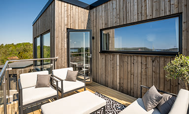 Modern takterrass med ljus träfasad, vita lounge-möbler, stålräcke och speglande fönster.