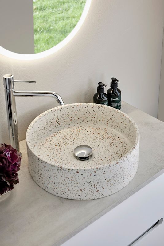 Rund vit terrazzohandfat med färgprickar på grå bänk, kromkran, svarta pumpflaskor och belyst spegel