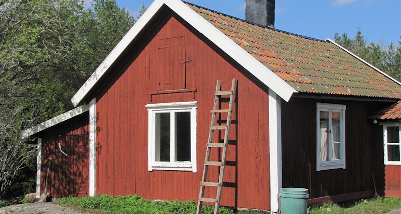 Röd trästuga med vita fönsterramar, mossigt tegeltak och trästege lutad mot väggen.