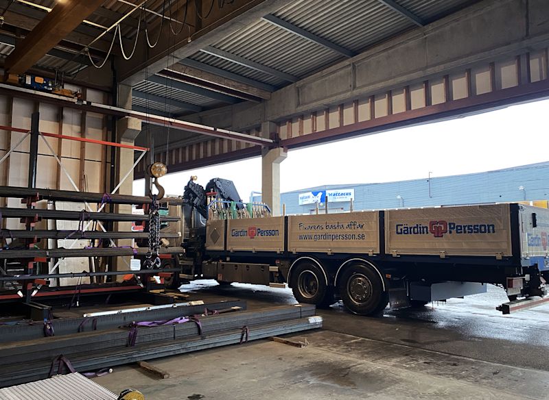 En lastbil med Gärdin & Persson-logotyp står inne i en lagerhall och är lastad med stålbalkar och byggmaterial.