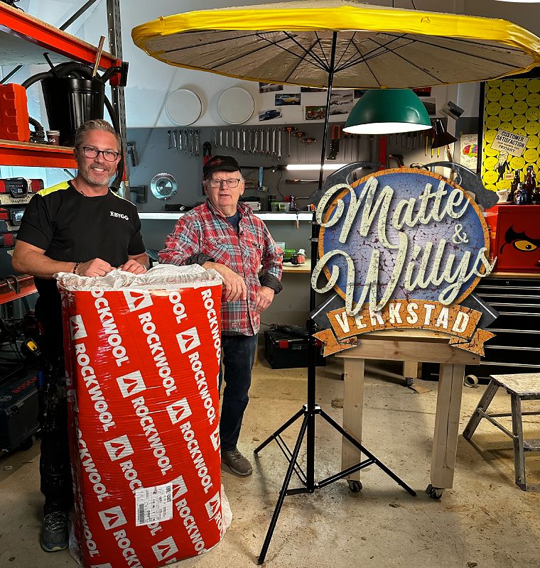 Två personer i verkstad med rött Rockwool-paket, gult paraply, skylt "Matte & Willys Verkstad"