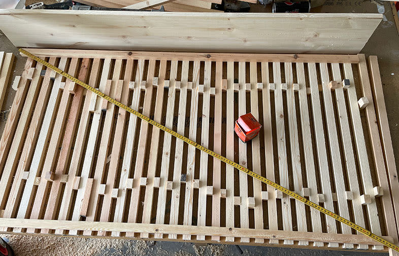 Ljus träram med parallella ribbor, gul tumstock diagonalt och rött måttband ovanpå i verkstad