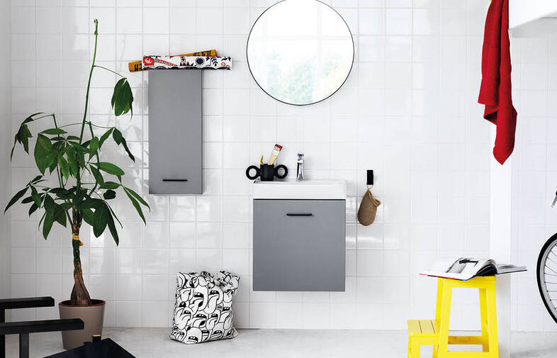 Ljust badrum med vita kakelväggar, grå skåp, runt spegel, grön växt, rött badlakan och gul pall.