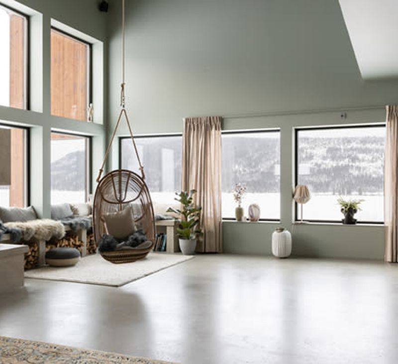 Ljust rum med gröna väggar, stora fönster mot snölandskap, rottinghängstol och växter.