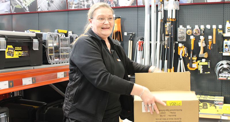 Person i svart öppnar kartong vid hylla med svarta och orange verktyg i butik.