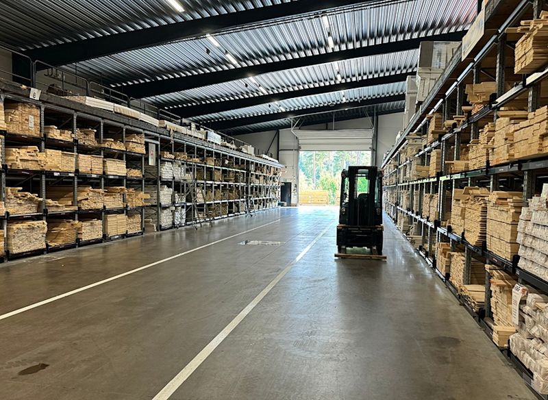 Lång lagerhall med metallhyllor fyllda av träbrädor och en gaffeltruck i gången