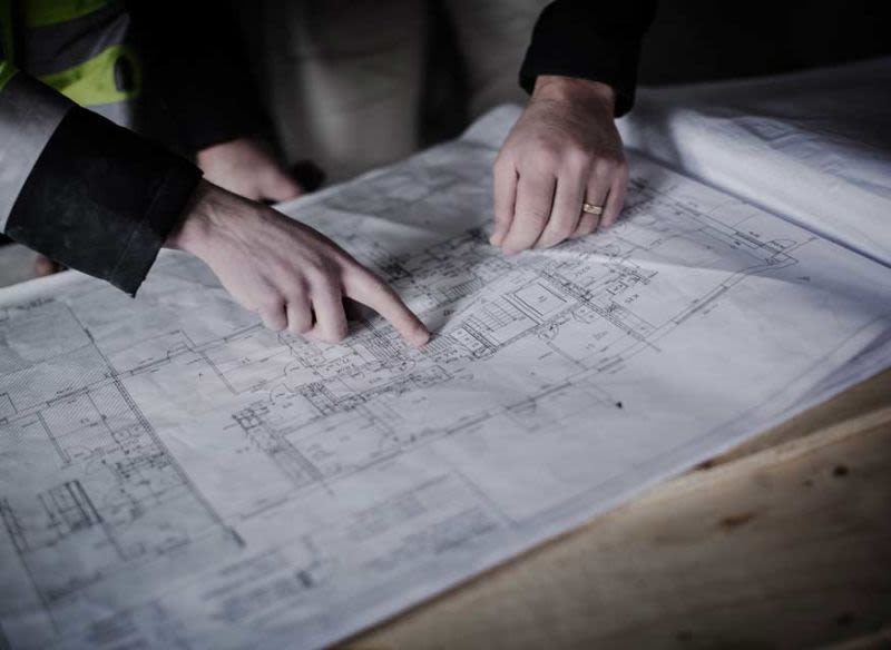 Två händer pekar på vit byggritning på träbord, svart ärm och gul reflexväst syns.