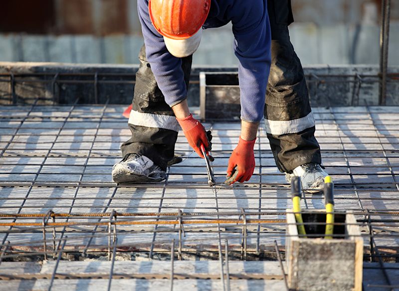 Byggarbetare i orange hjälm och röda handskar binder armeringsjärn på betonggolv.