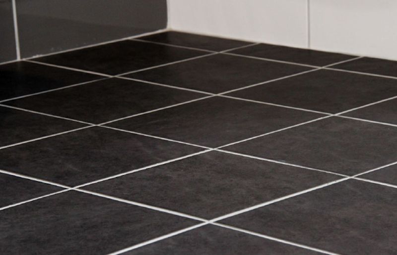 Mörka kvadratiska golvplattor i svartgrå ton med vita fogar mot vit vägg.