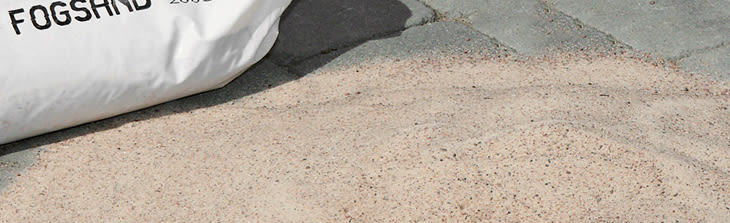 Vit säck märkt "FOGSAND" på grå gatstensyta med utspilld beige fogsand runt.